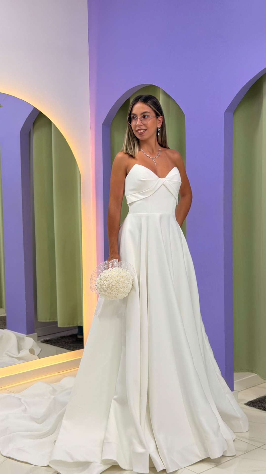 Vestido Celine Noiva Vestido de noiva branco com corpete tomara que caia e saia ampla, bouquet de flores, mulher com joias e óculos em fundo com arcos violetas.