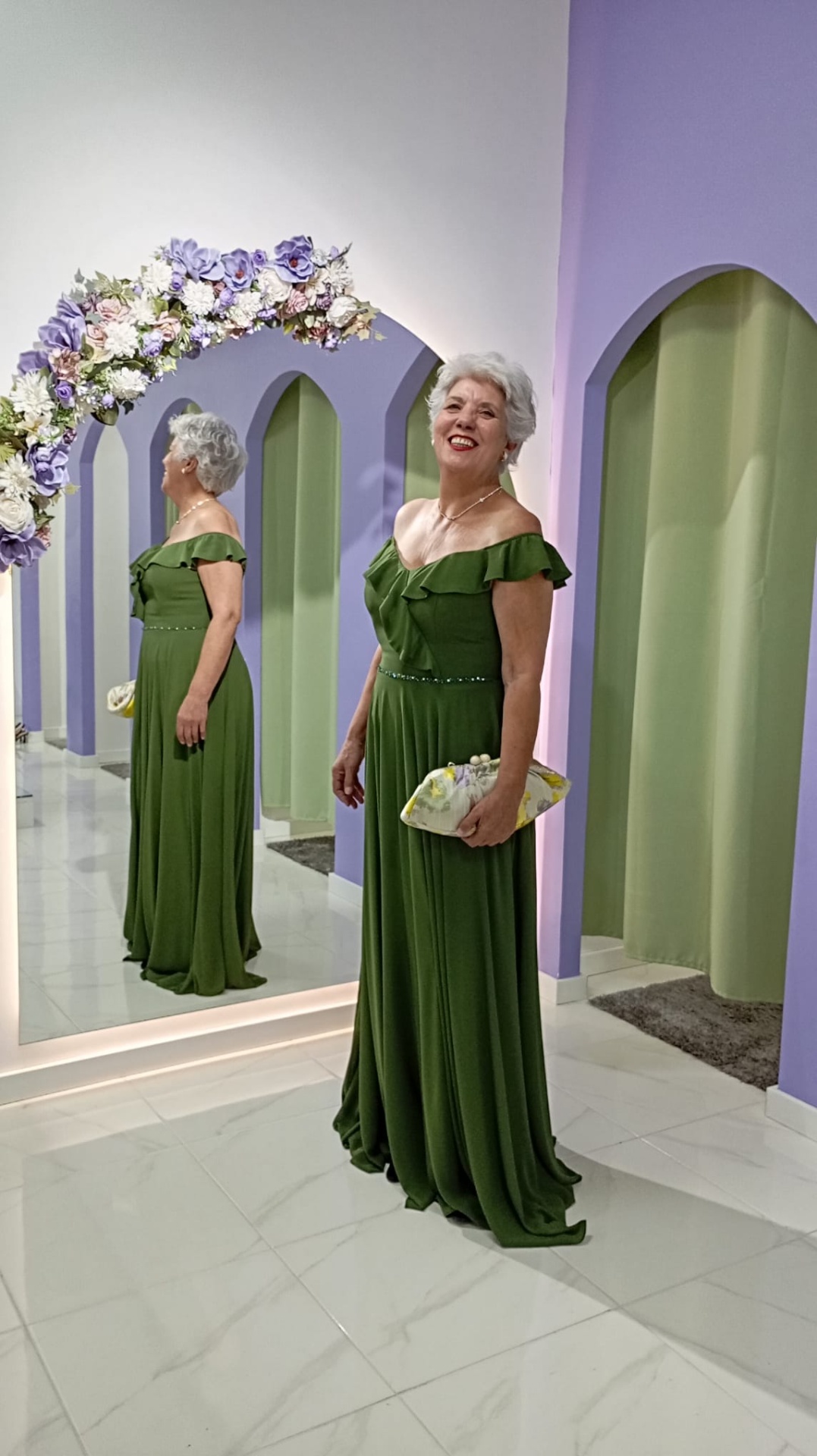 Vestido Nataly Mulher com vestido verde comprido em ambiente com paredes lilás e espelho decorado com flores