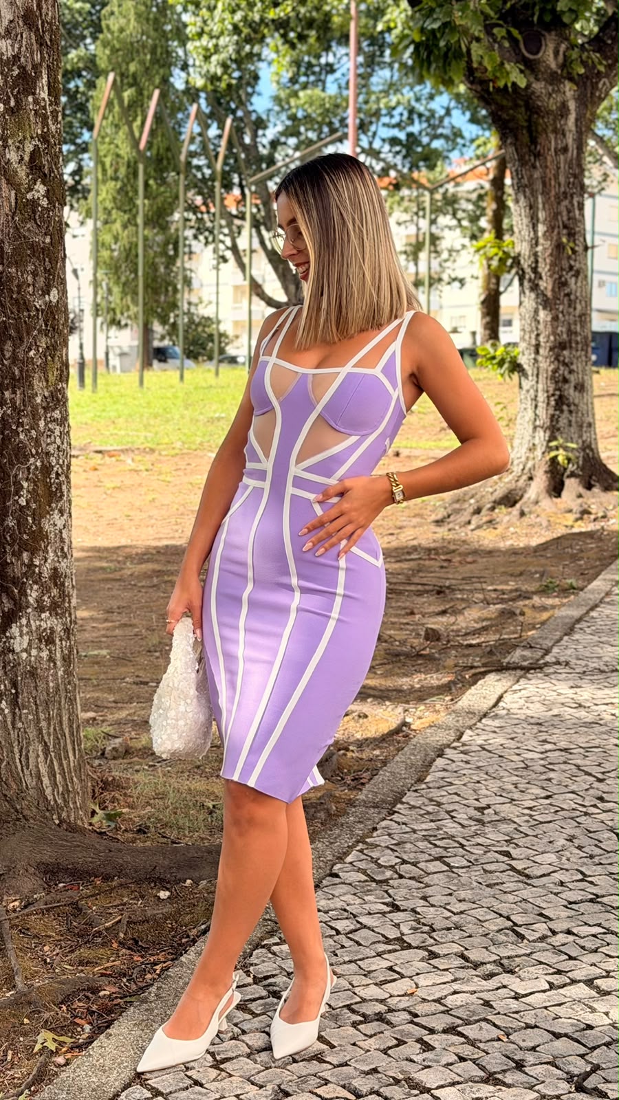 Vestido ALFAZEMA lilás Mulher de vestido lilás justo com detalhes brancos e painéis transparentes, sapatos brancos, bolsa branca, em ambiente exterior com árvores.