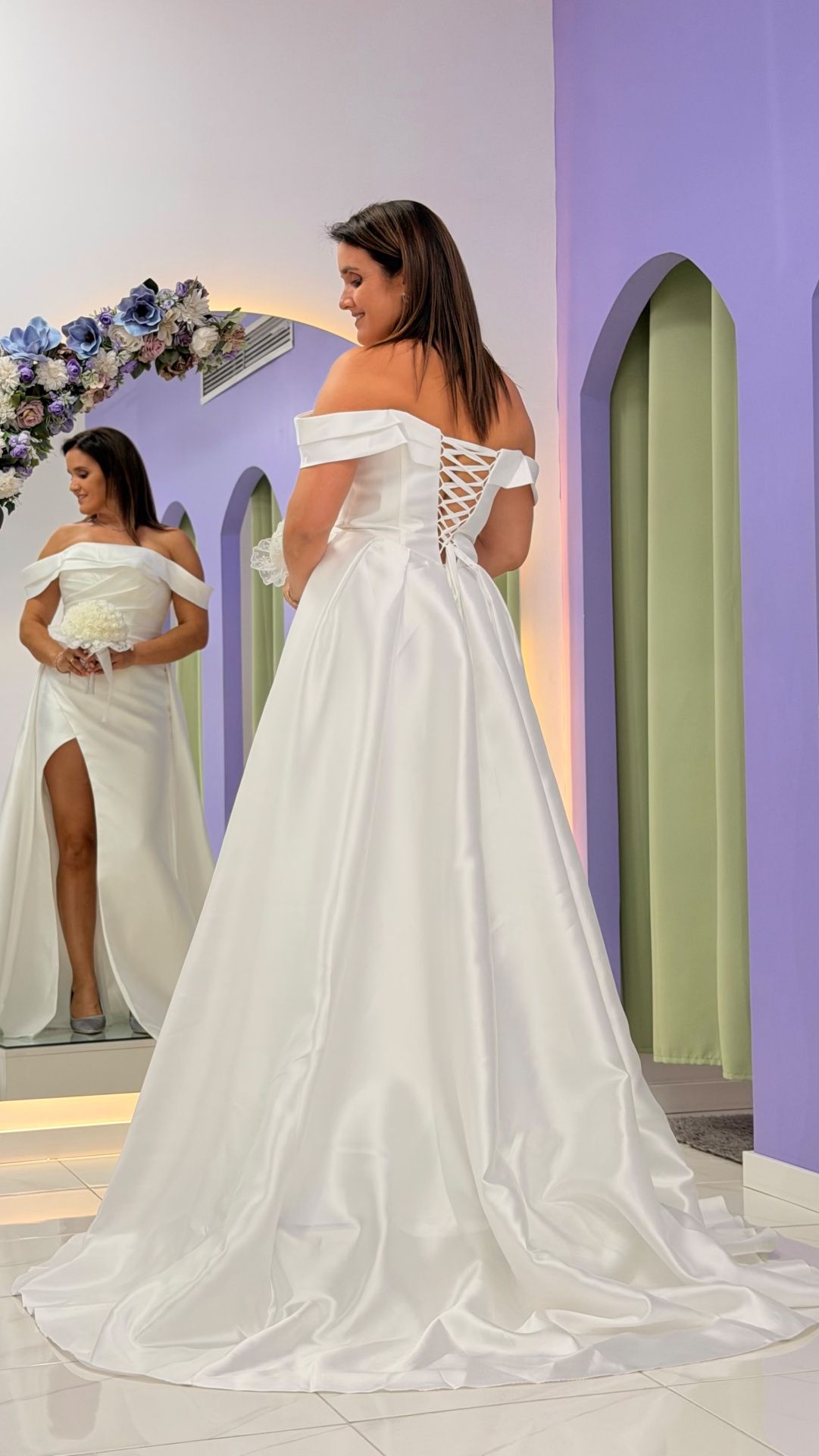Vestido Aurora Noiva Vestido de noiva branco com ombros descobertos e amarração nas costas, refletido num espelho com decoração floral e parede lilás com arcos verdes.