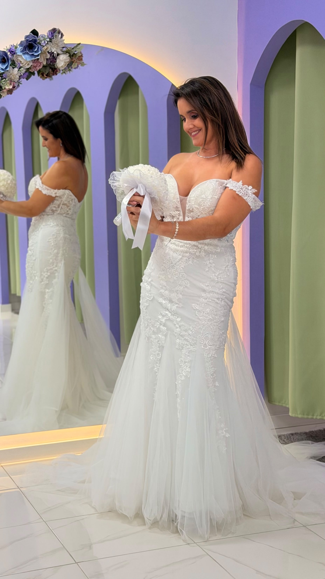 Vestido Wedding Ilusion Mulher com vestido de noiva branco rendado e bouquet em ambiente interior com parede lilás e verde