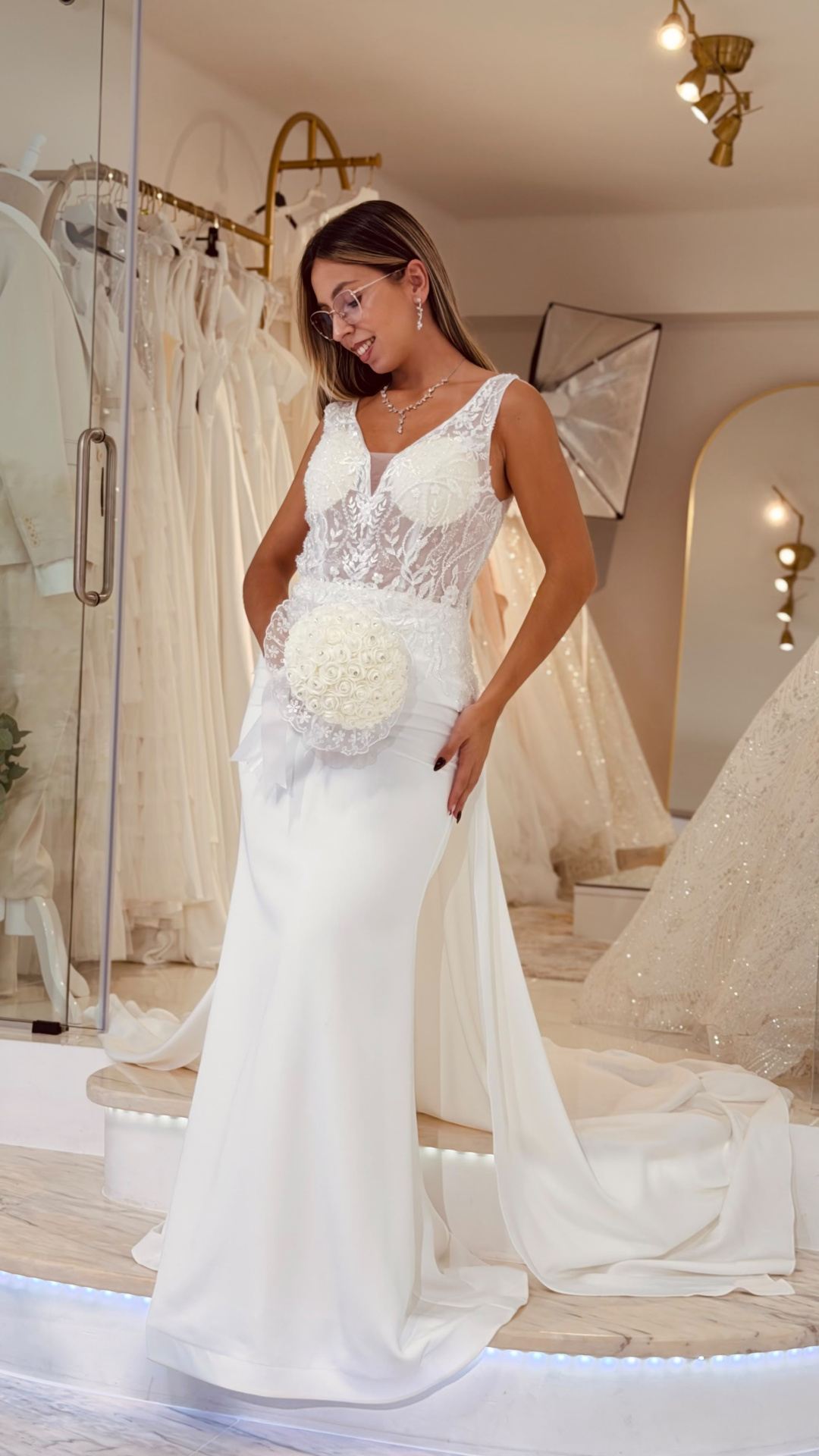 Vestido Denielle Noiva Mulher com vestido de noiva branco com renda e bouquet redondo, em loja de vestidos de noiva.