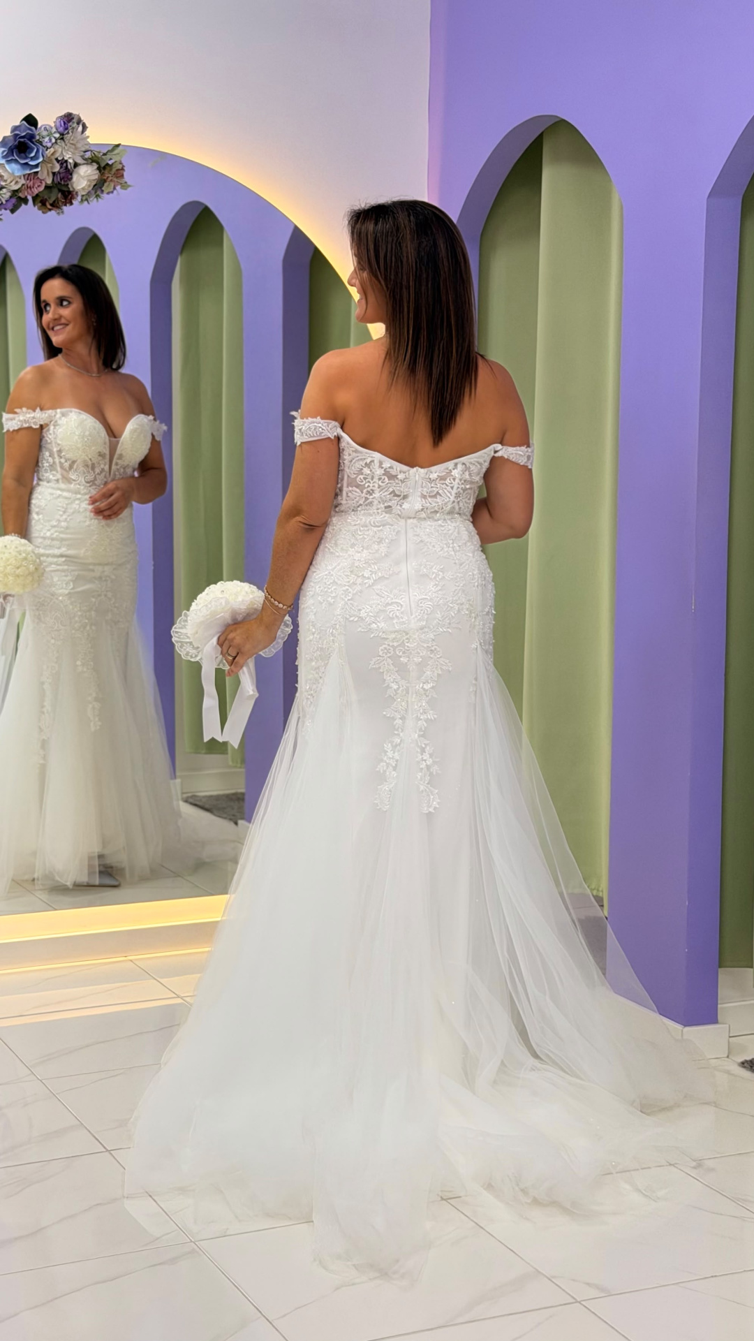 Vestido Wedding Ilusion Mulher com vestido de noiva branco com renda e tule diante de espelho e paredes lilás e verdes