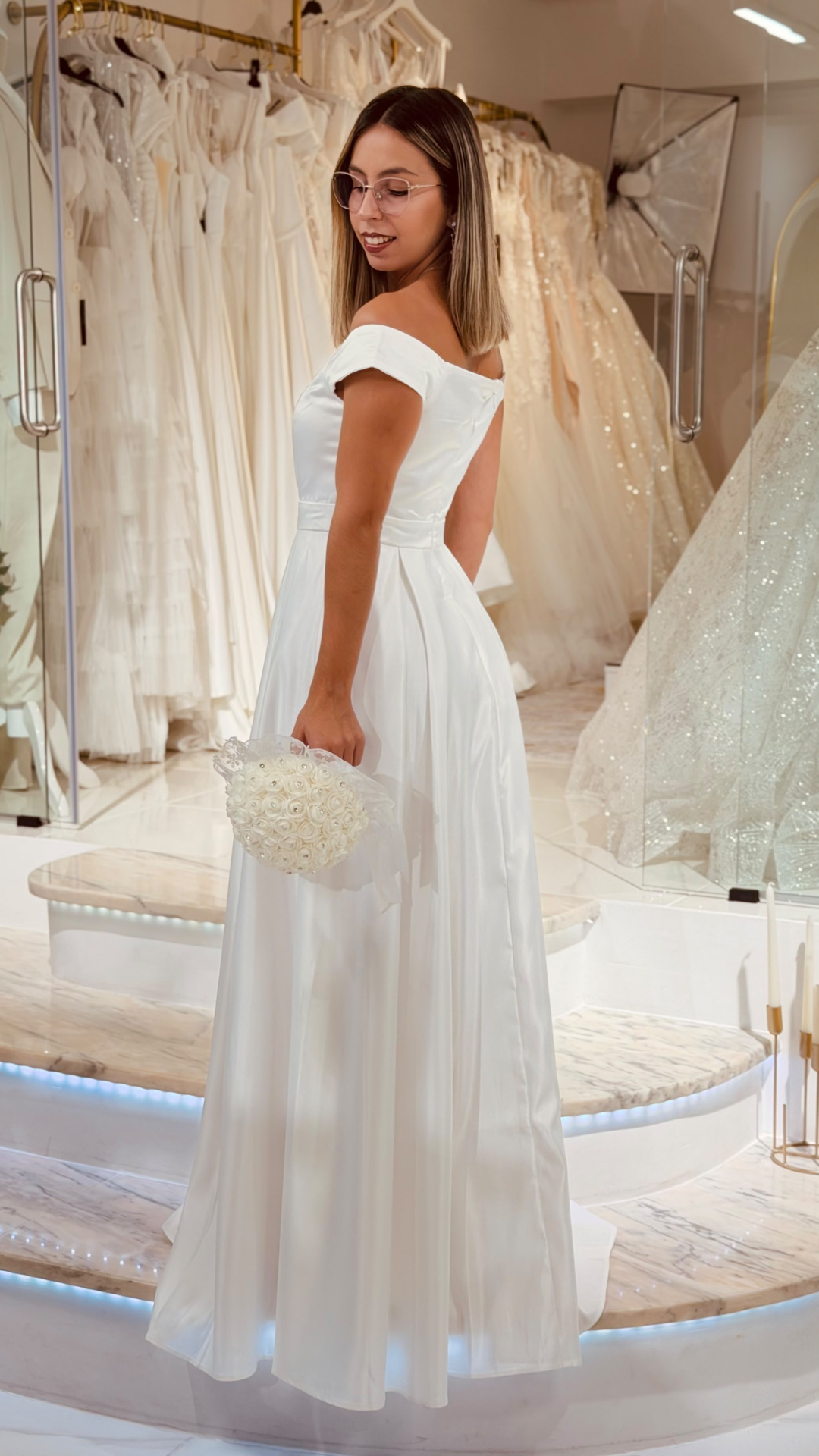 Vestido Santa Noiva Vestido de noiva branco de tecido acetinado com corte ombro a ombro e bouquet branco em loja