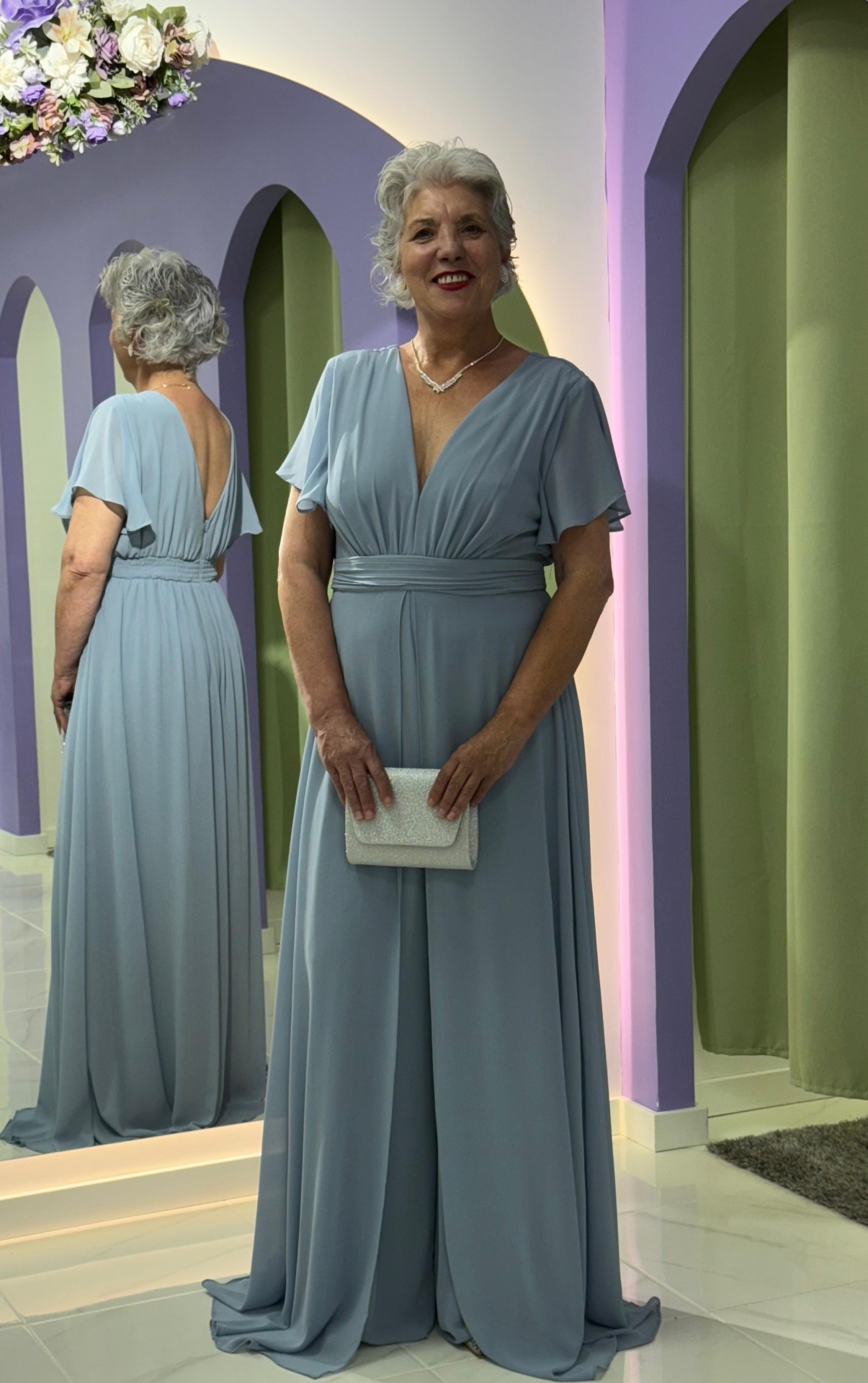 Macacão/Vestido Plus Size Mulher com vestido comprido azul claro com decote em V, segurando mala branca, em frente a espelho
