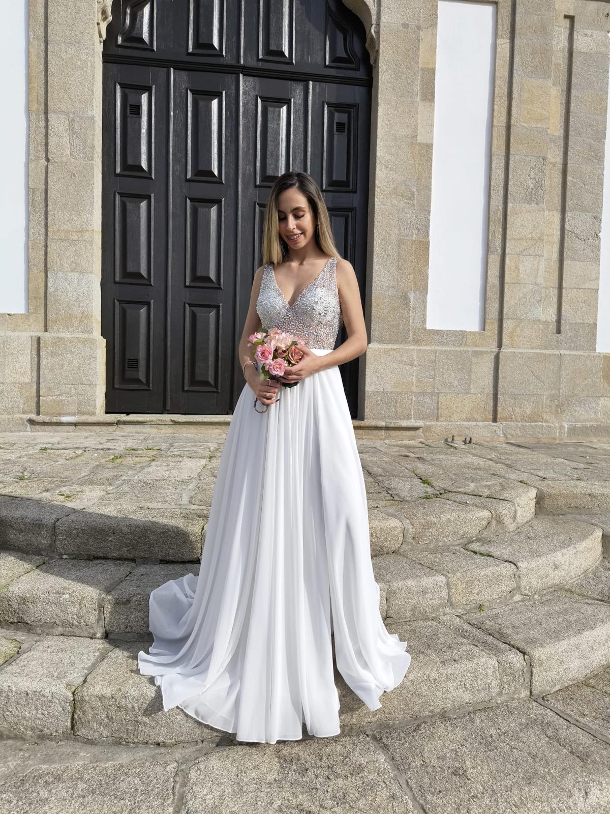 Vestido Simplicidade Serena Mulher com vestido de noiva branco e lantejoulas prateadas segurando flores cor-de-rosa em degraus de pedra