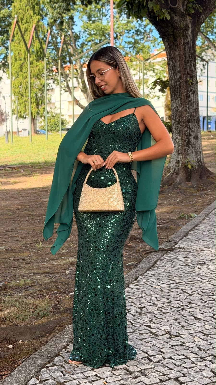 Vestido Milene Lantejoulas Mulher com vestido verde de lantejoulas e lenço, segurando mala dourada em exterior