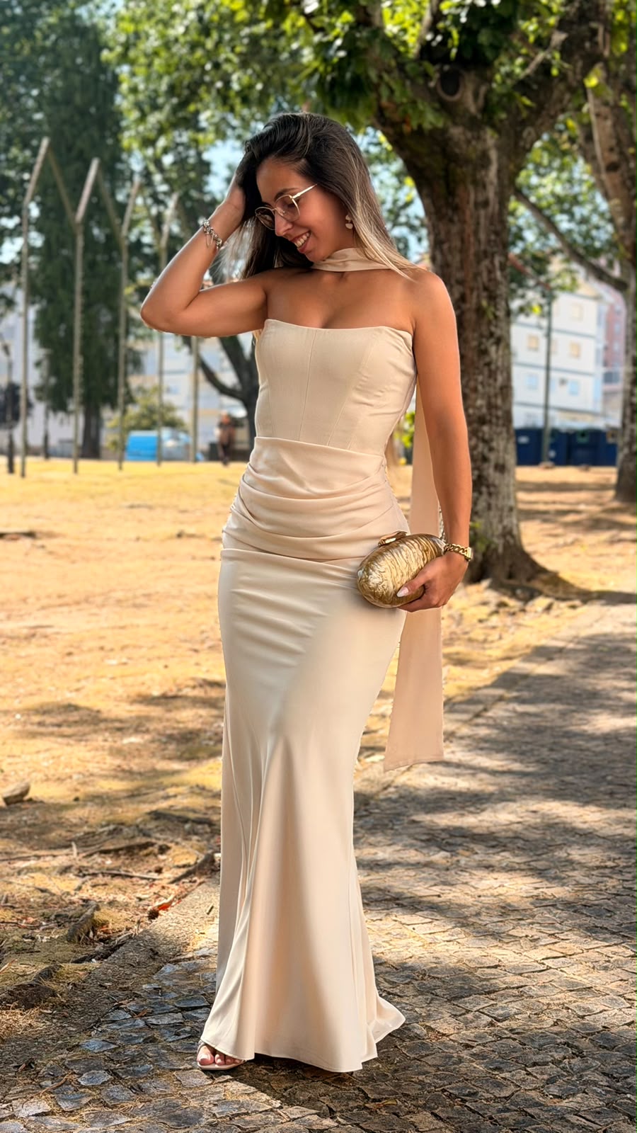 Vestido Romana Mulher com vestido longo crema e clutch dourada ao ar livre
