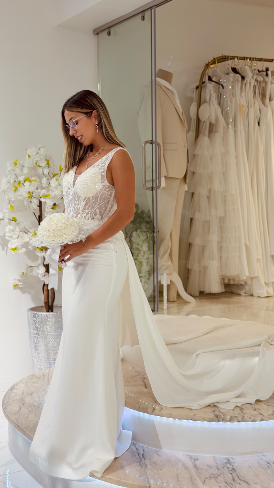 Vestido Denielle Noiva Vestido de noiva branco rendado com buquê e cauda longa numa loja.
