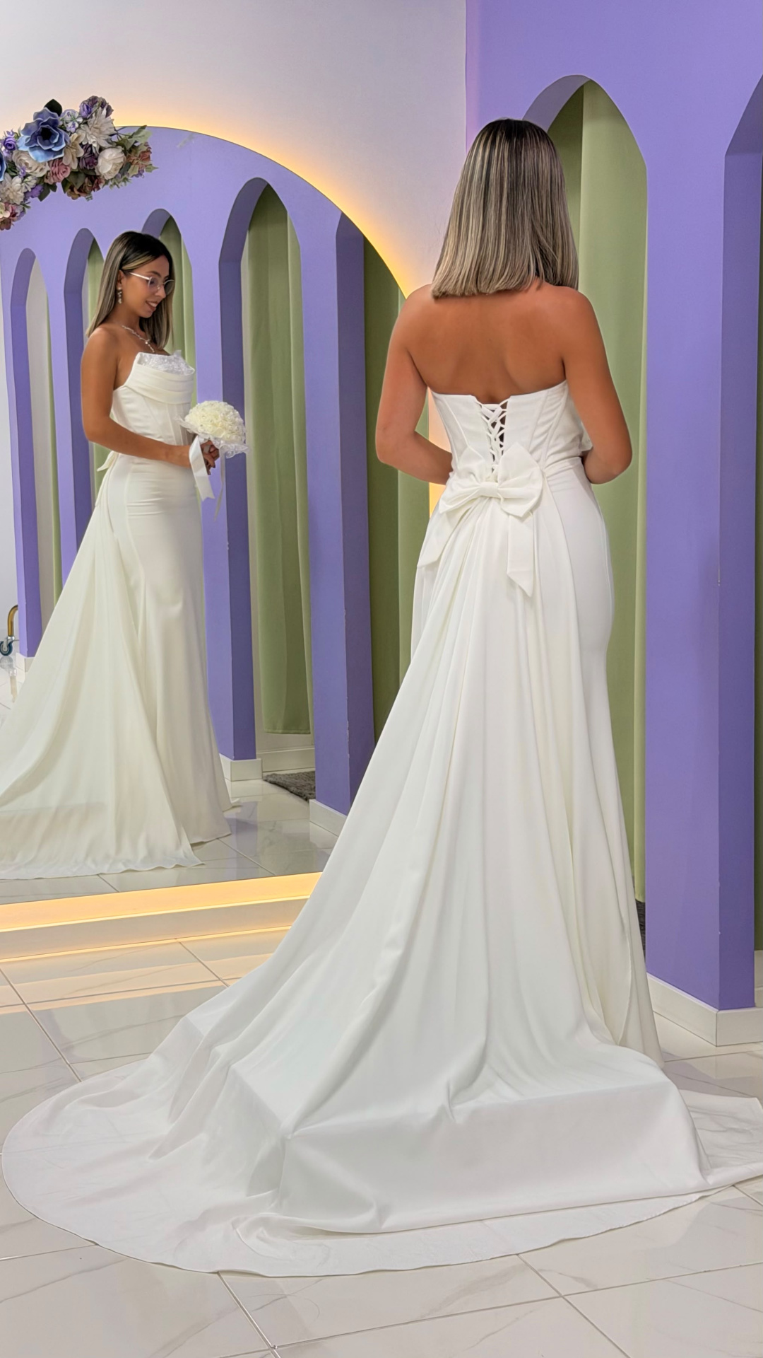 Vestido Éden Noiva Vestido de noiva branco com laço e cauda em ambiente com paredes lilás
