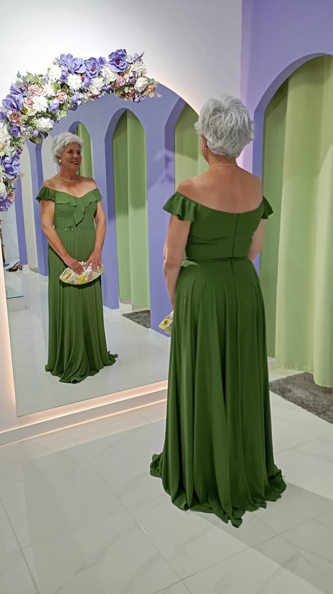 Vestido Nataly Senhora com vestido longo verde e carteira dourada em frente a espelho decorado com flores.