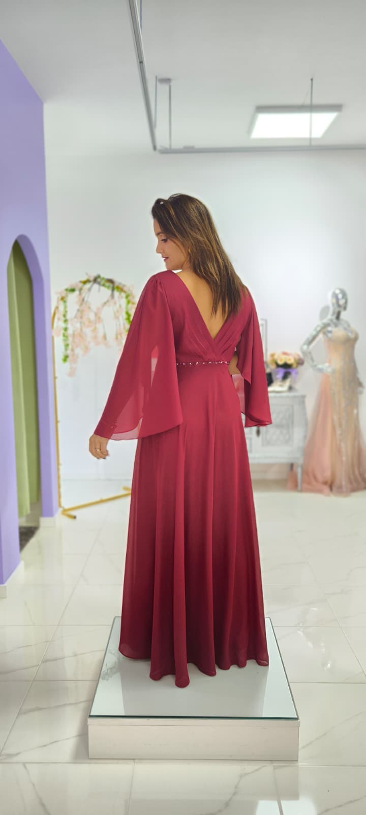 Vestido Leonor c/ Capa Plus Size Vestido longo vinho com mangas largas e decote V nas costas numa sala clara com chão de mármore.