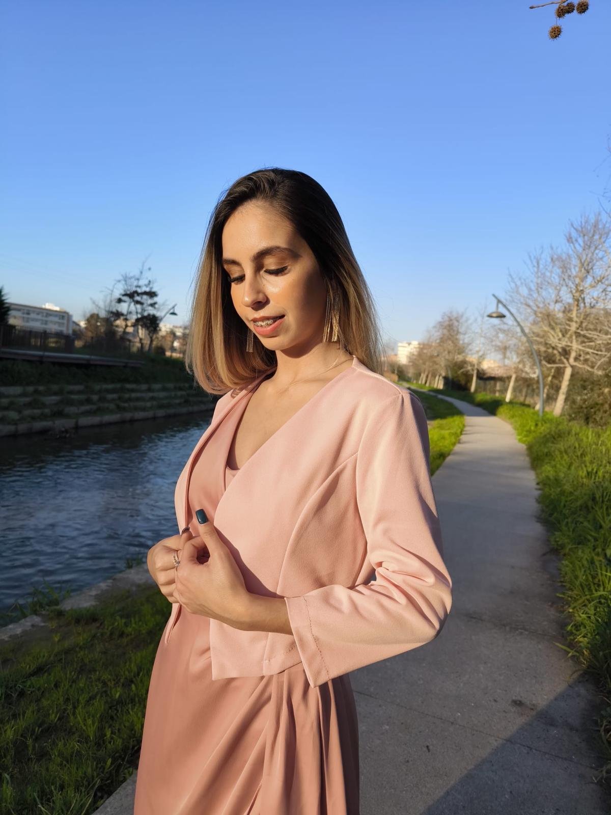 Bolero Simply Mulher com vestido rosa claro e casaco curto rosa junto a um rio num dia ensolarado