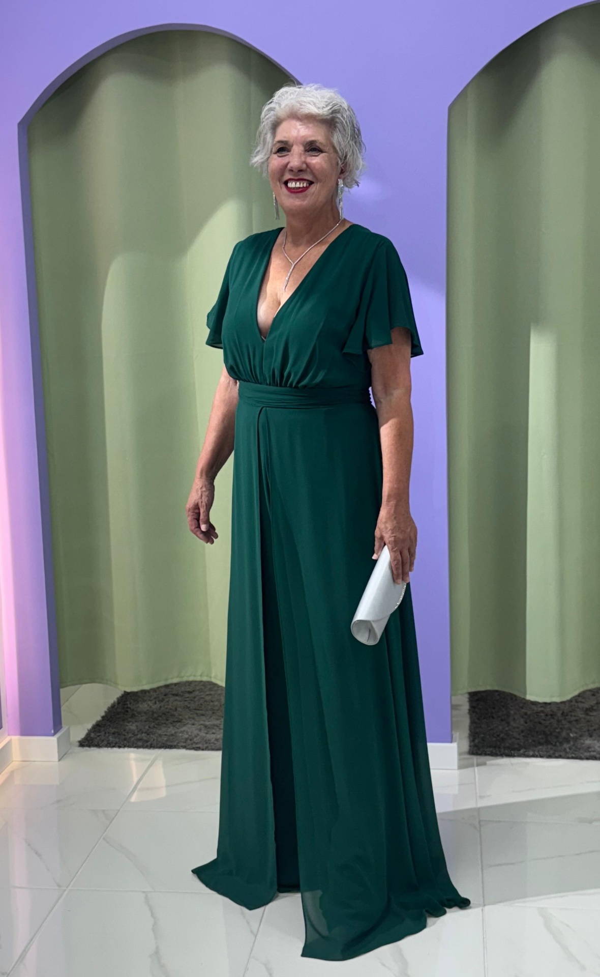 Macacão/Vestido Plus Size Mulher com vestido verde escuro longo e carteira branca em interior com paredes lilás.