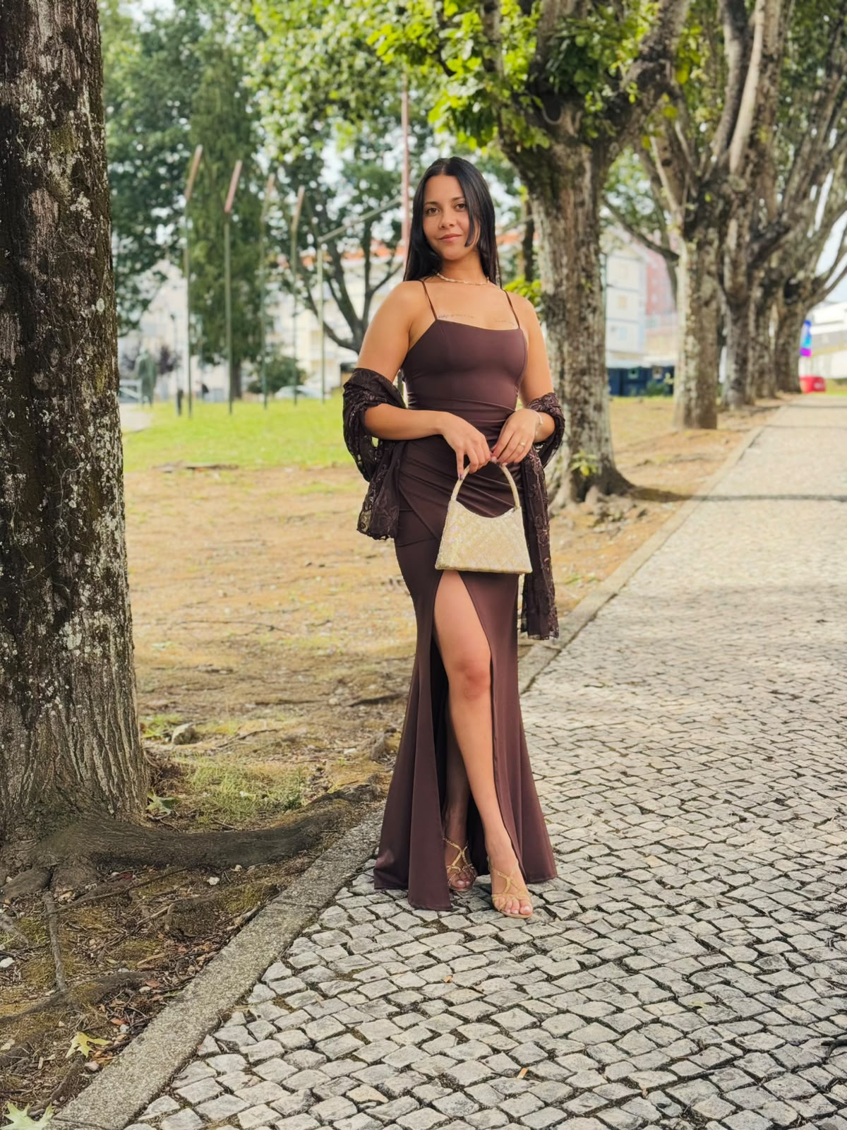 Vestido HUNGARO Mulher com vestido longo castanho e mala pequena bege em calçada com árvores