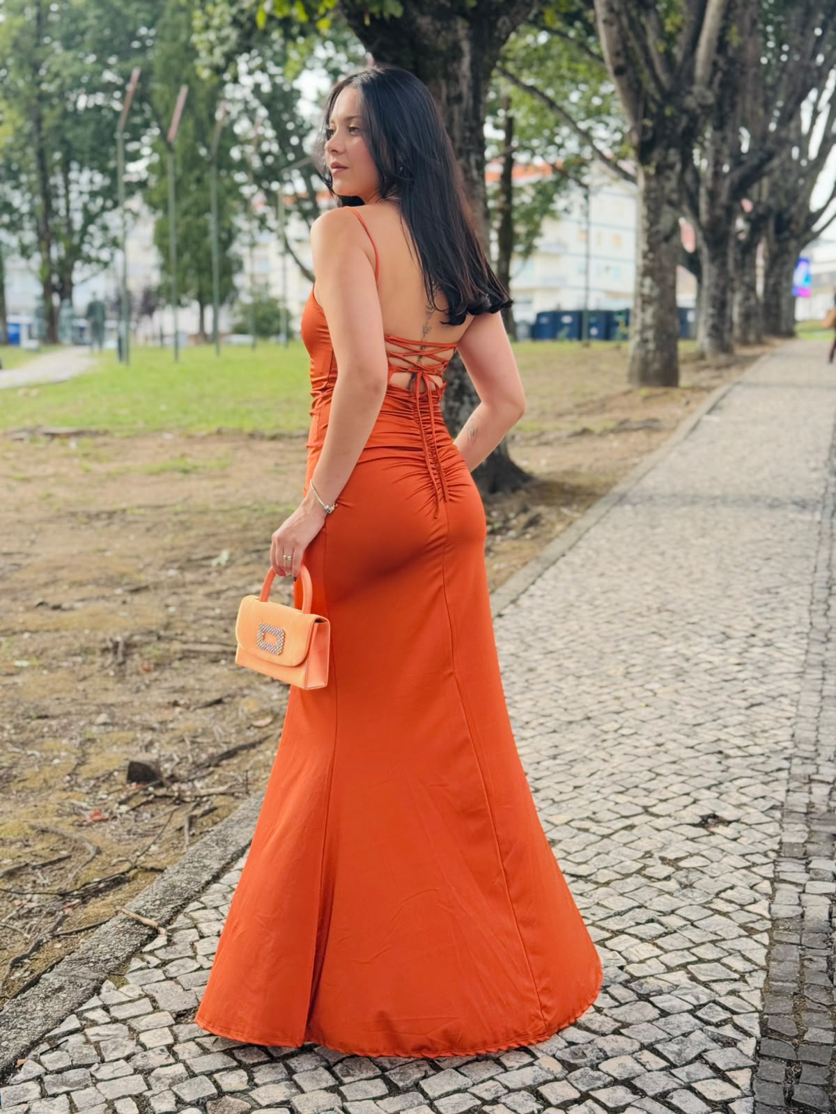 Vestido HUNGARO Modelo usa vestido longo laranja de cetim com alças finas e mala de mão laranja ao ar livre.