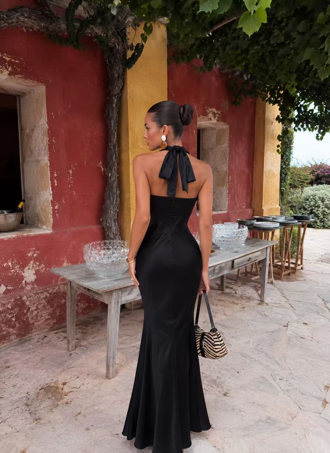 Vestido TULIPA Mulher de costas com vestido preto longo e laço, segurando mala zebra, em exterior com parede vermelha e amarela