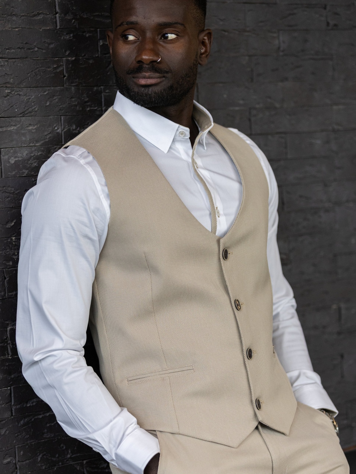 Colete SMK Golden Wear Homem de pele escura com colete bege e camisa branca encostado numa parede preta