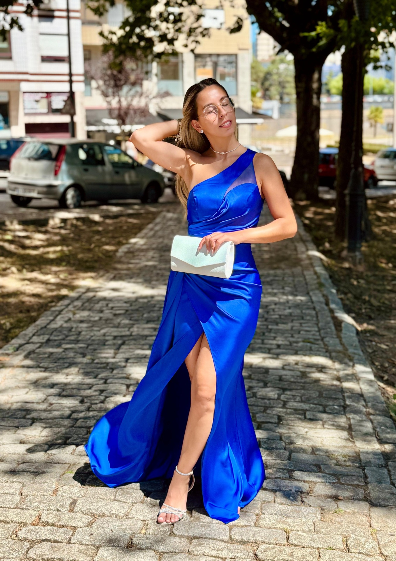 Vestido Mayse Mulher com vestido azul royal brilhante, clutch branca e sandálias prateadas numa rua de pedra com carros e árvores