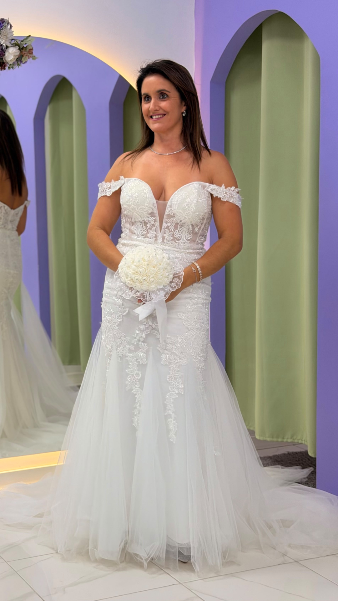 Vestido Wedding Ilusion Mulher a usar vestido de noiva branco com detalhes de renda e tule, segurando bouquet de rosas brancas, junto a fundo com arcos lilás e cortinas verde-claras.