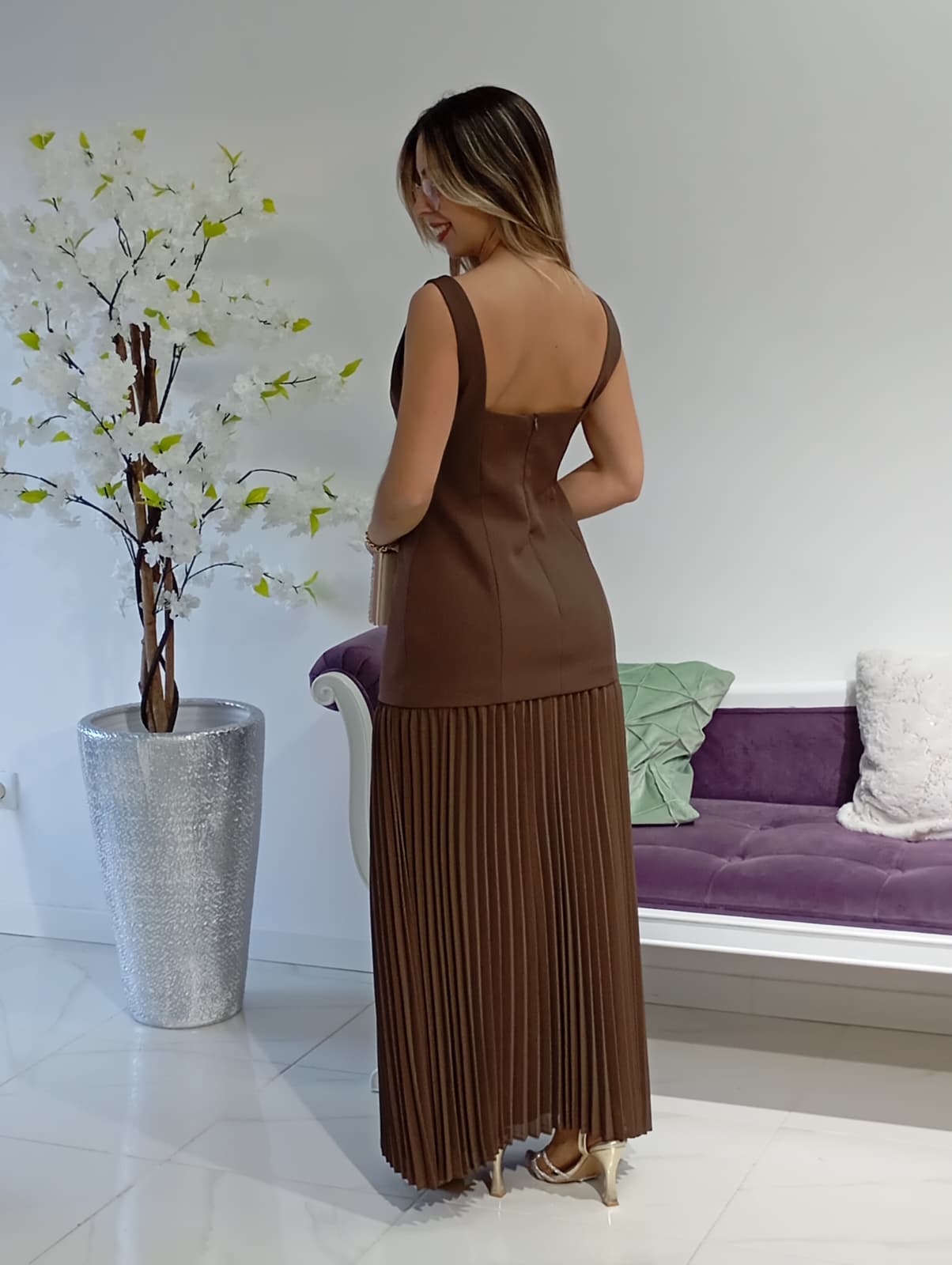 Vestido Vera&Lucy Vestido castanho com saia plissada, visto por trás, em ambiente interior com vaso prateado e sofá roxo