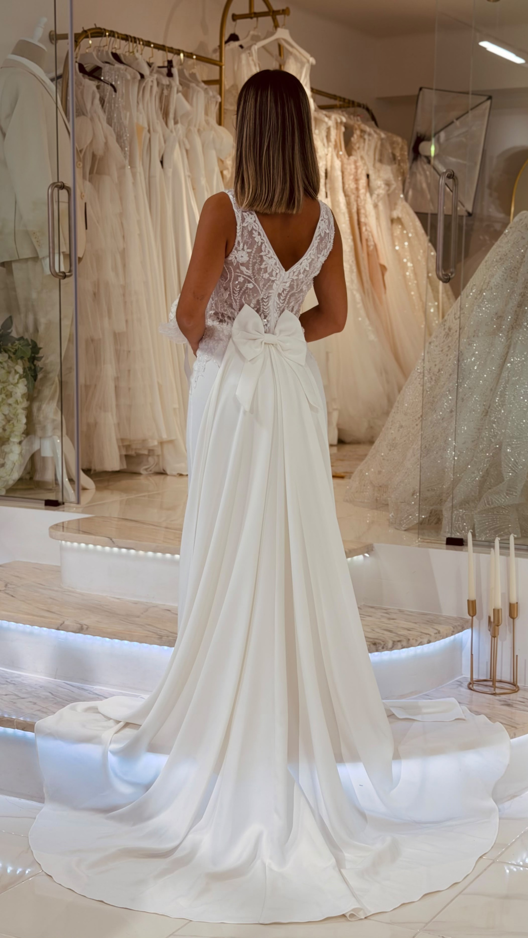 Vestido Denielle Noiva Vestido de noiva branco com laço e renda, visto por detrás, numa loja de vestidos.