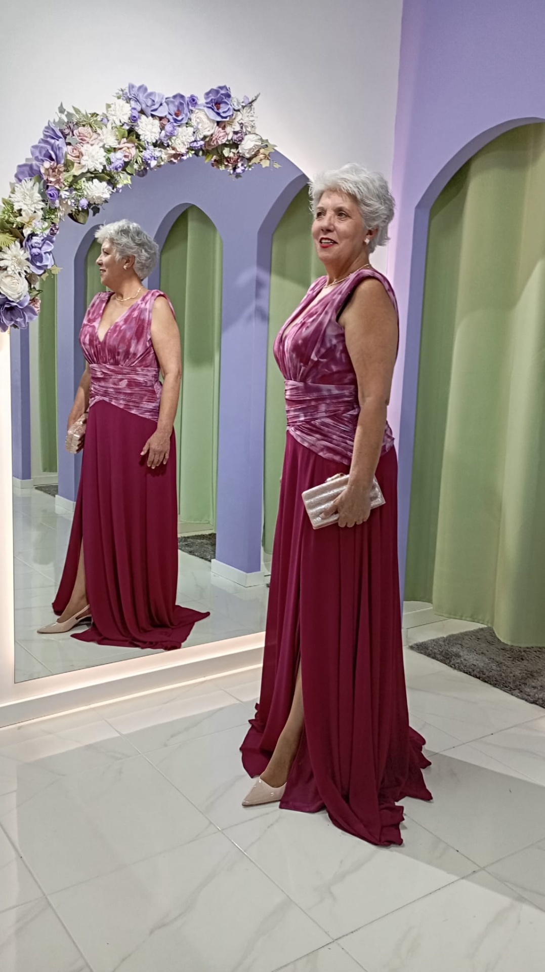 Vestido Andore Plus Size Mulher com vestido longo vinho e rosa em frente a espelho com decoração floral e cortinas verdes.