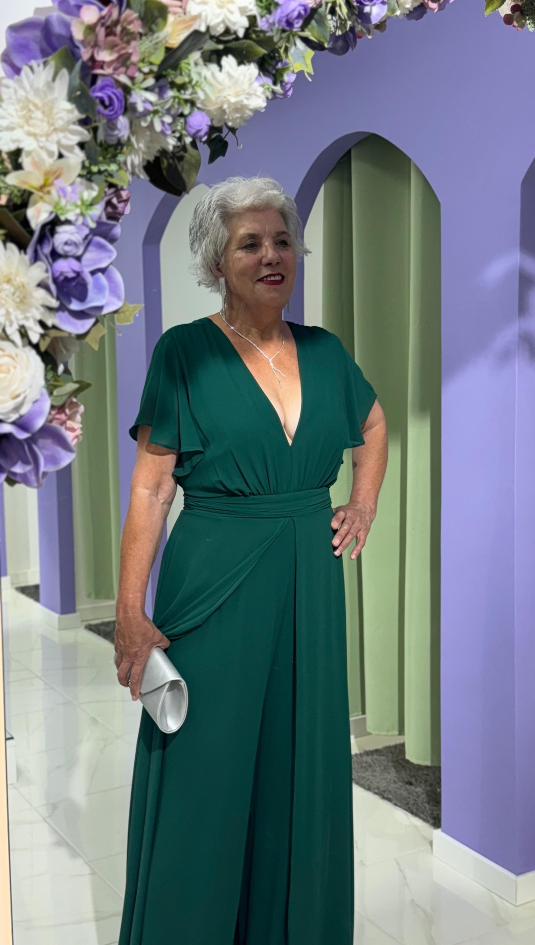 Macacão/Vestido Plus Size Mulher com vestido longo verde escuro e carteira branca em ambiente interno com paredes lilás e verde