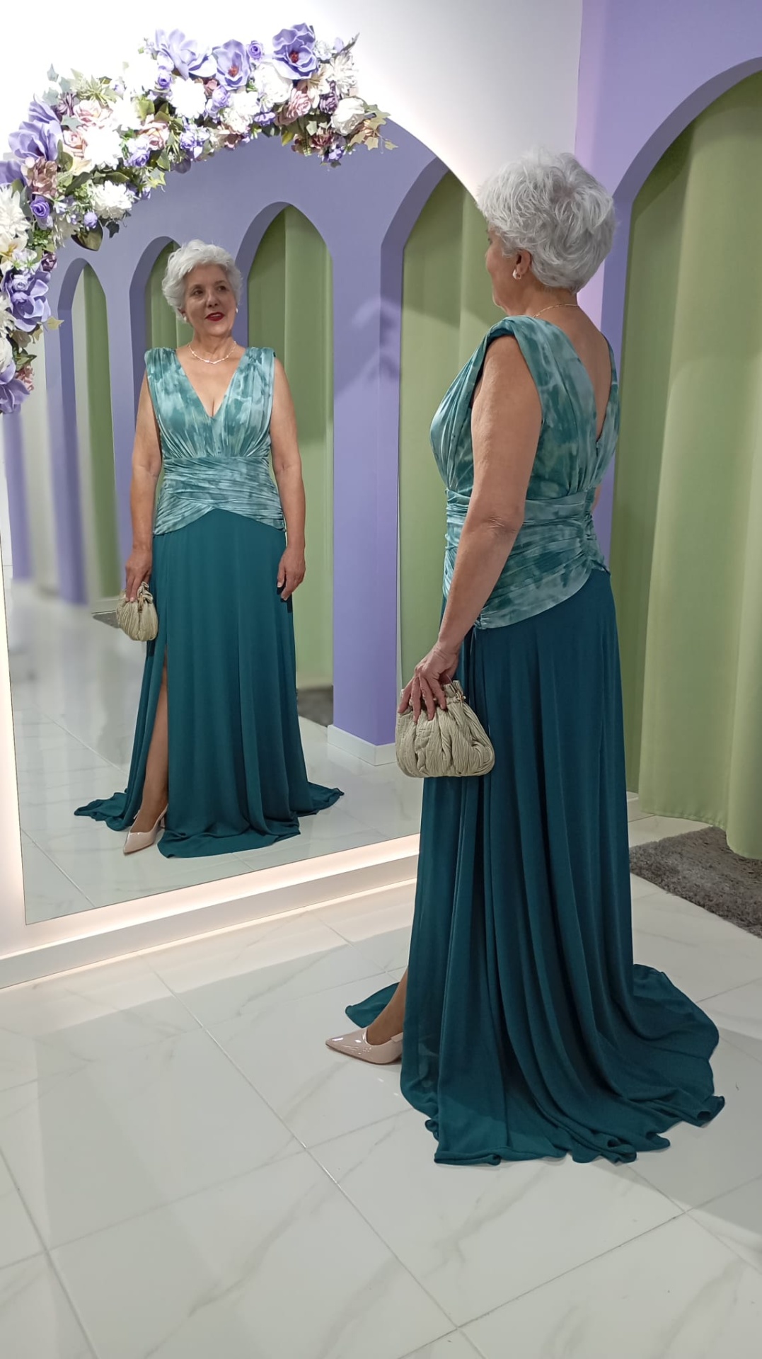 Vestido Andore Plus Size Mulher com vestido comprido azul esverdeado e parte superior azul claro franzida, segurando mala bege, de frente para espelho num espaço com paredes lilases e arco de flores