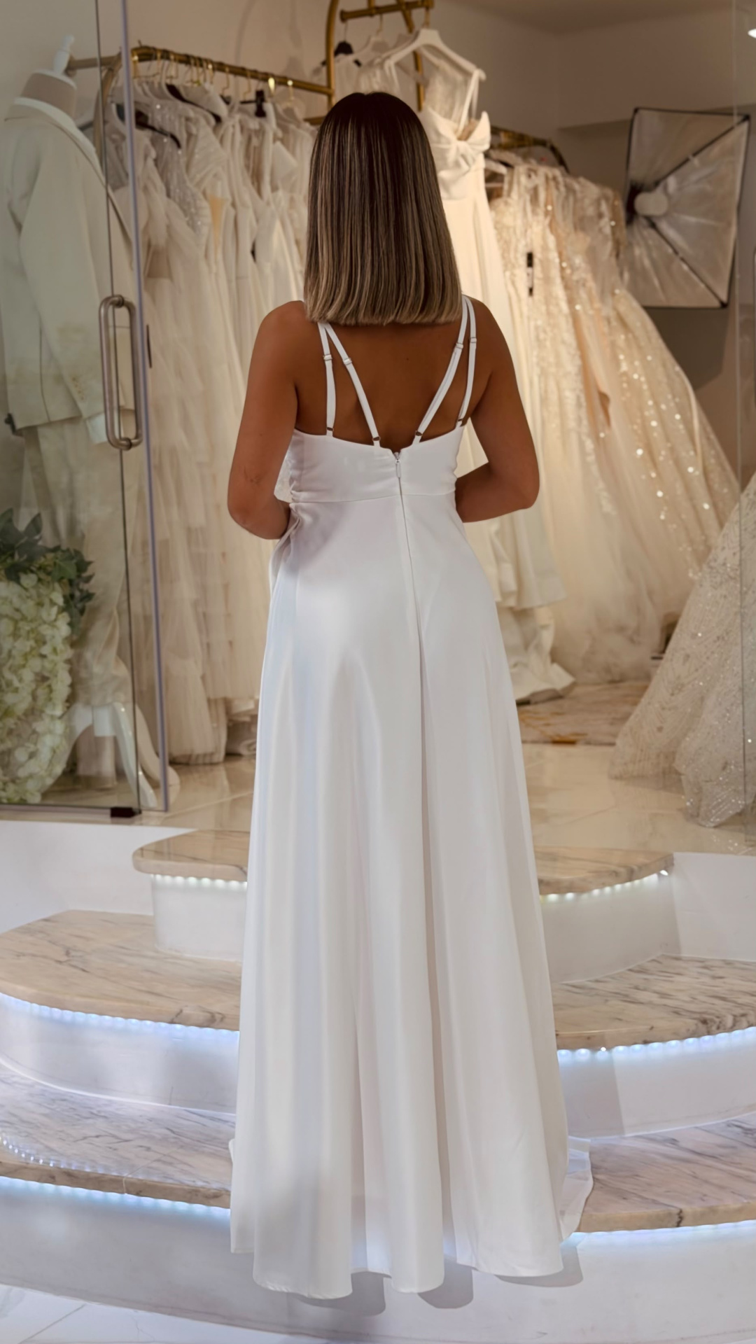 Vestido Muse Noiva Mulher de costas a usar vestido branco longo com alças duplas, numa loja de vestidos de noiva.