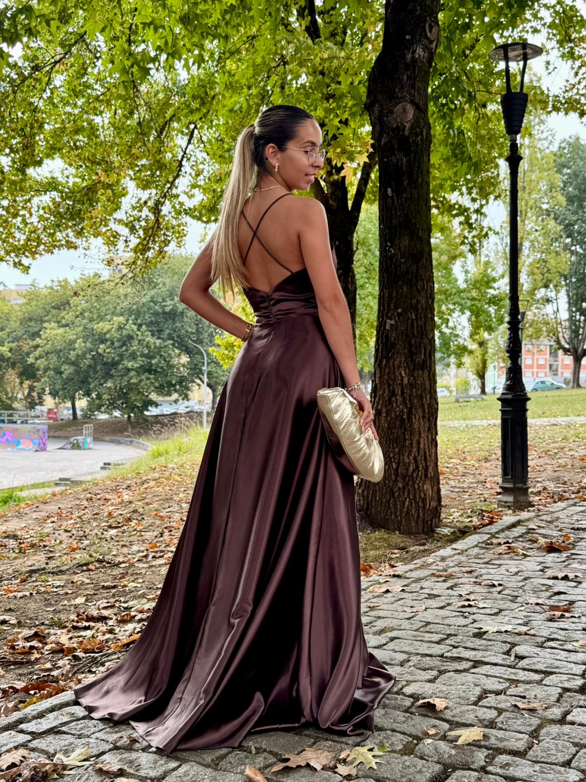 Vestido Doriane Mulher de costas com vestido longo castanho de cetim e mala dourada ao ar livre