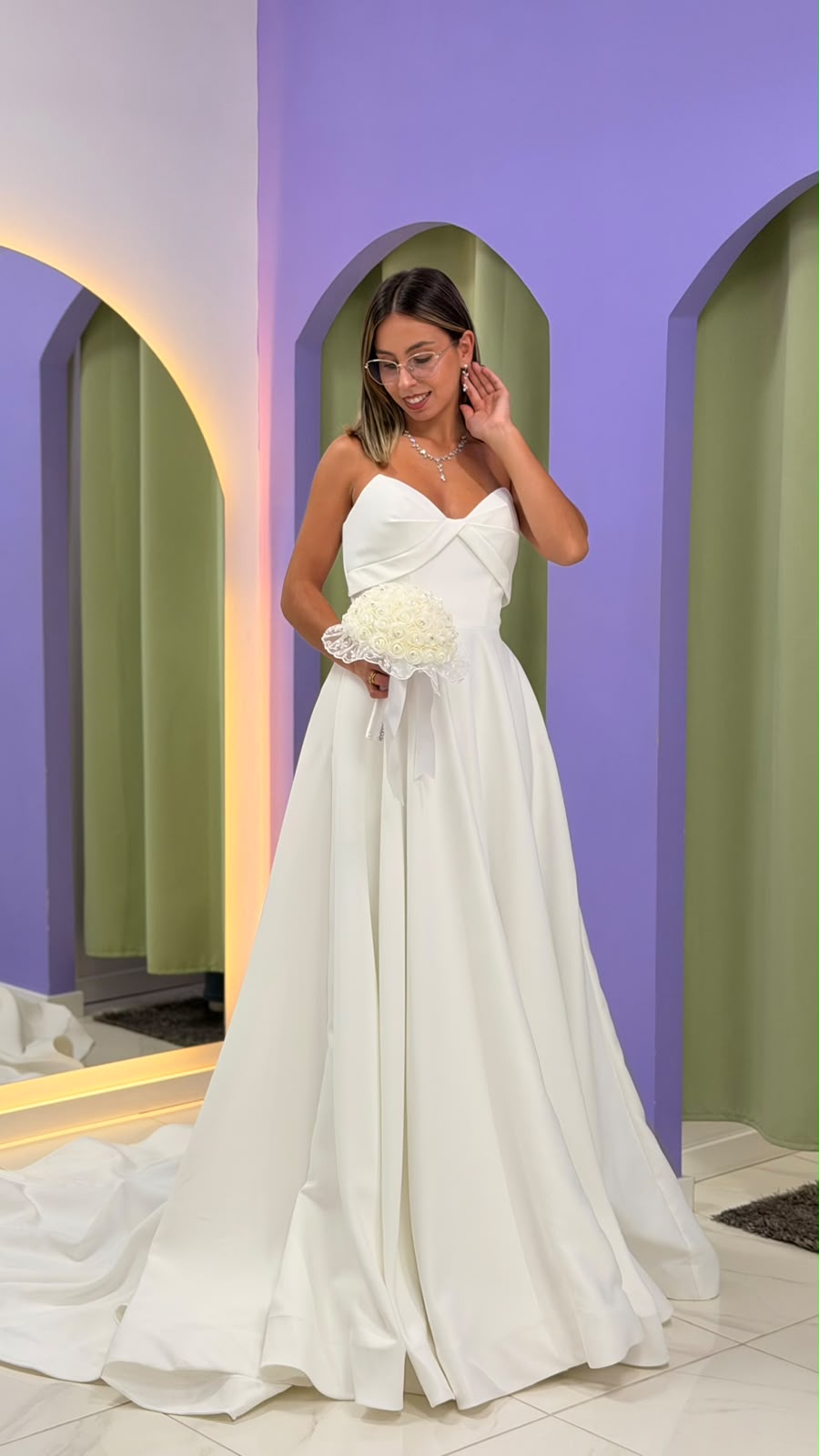Vestido Celine Noiva Jovem mulher com vestido de noiva branco segurando ramo de flores, com parede roxa e cortinas verdes ao fundo