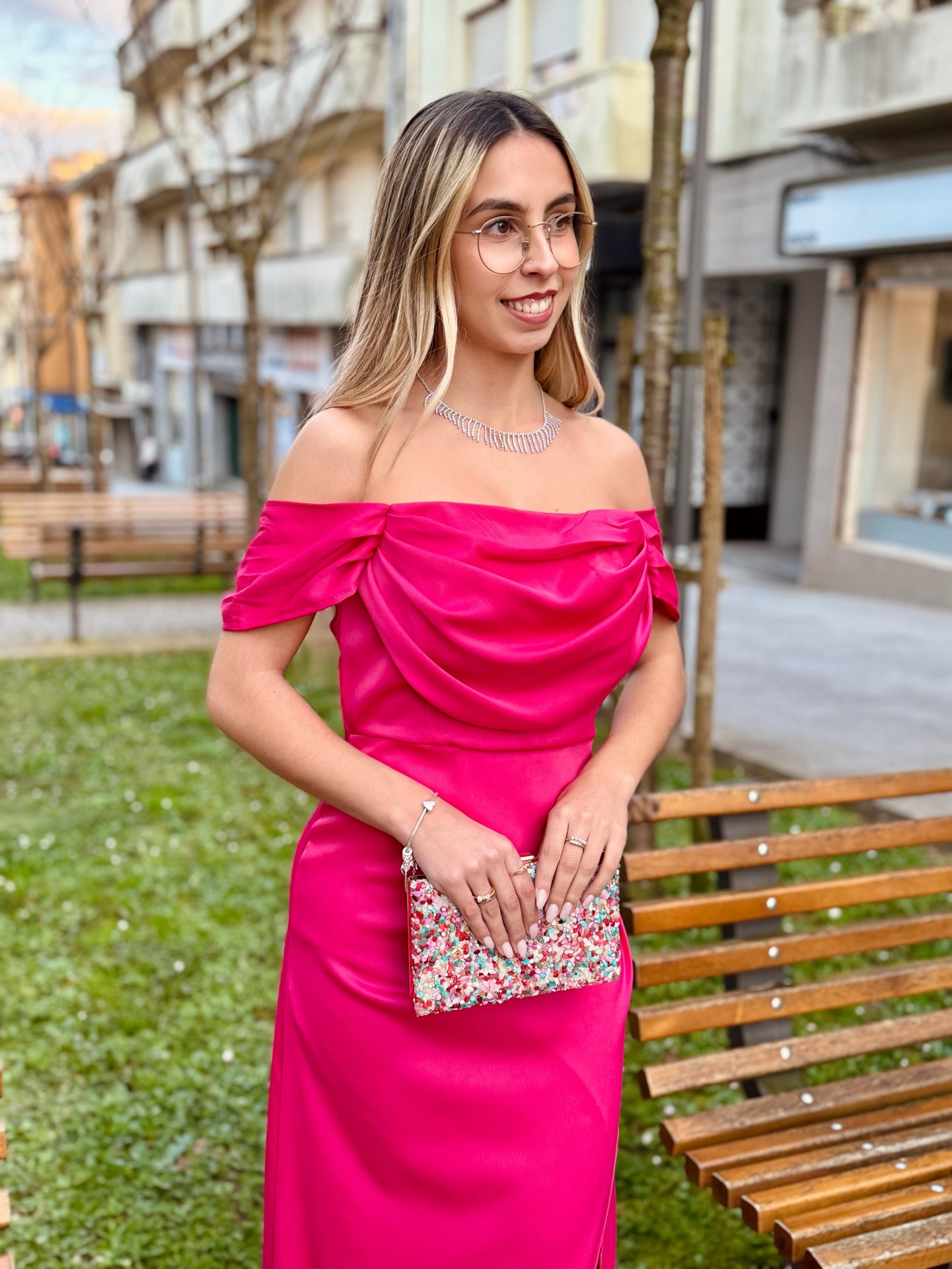 Vestido Sony Mulher com vestido rosa cetim decote ombro a ombro segurando carteira estampada