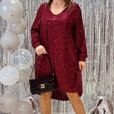 Mulher com vestido bordô de lantejoulas, mala preta e sapatos de salto junto a balões e cortinas prateadas