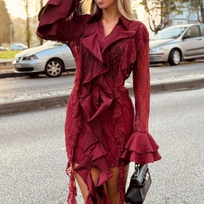 Mulher com vestido vinho de renda com folhos, botas pretas e mala preta numa rua urbana.
