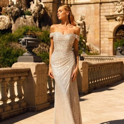 Vestido dourado longo brilhante ombro a ombro numa área exterior com arquitetura clássica.