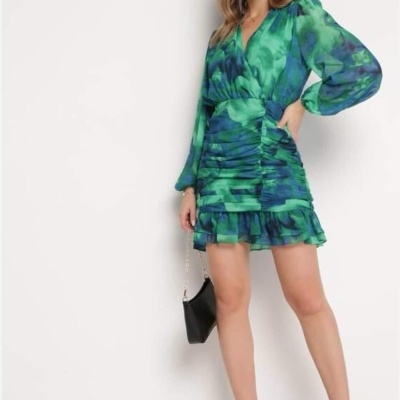 Vestido curto verde e azul com manga longa e franjas, mulher com bolsa preta e sandálias pretas de salto.