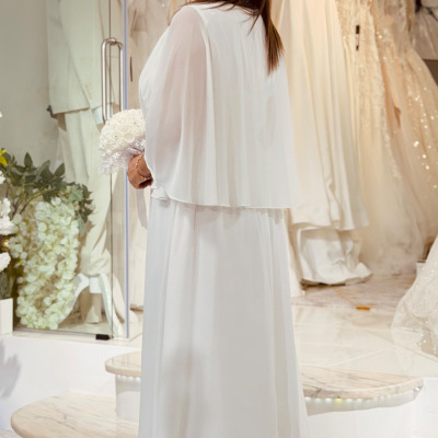 Mulher de costas a usar vestido de noiva branco com capa curta, segurando bouquet, em loja de vestidos de noiva.
