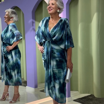 Vestido midi azul e verde com padrão abstrato, senhora sorridente diante de espelho e parede lilás
