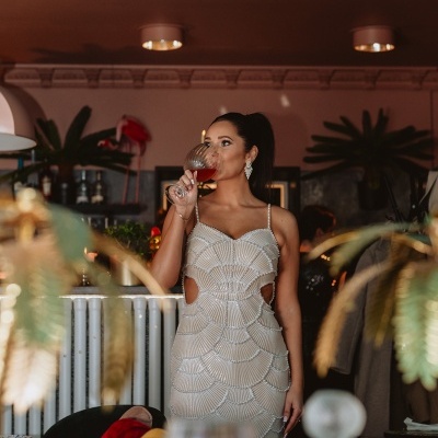 Mulher com vestido de festa branco com padrão de escamas a beber numa taça de vidro em restaurante moderno