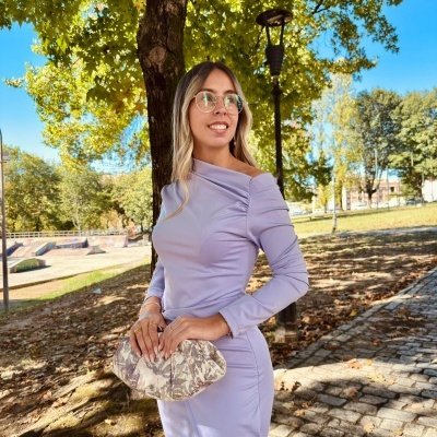 Mulher com vestido lilás e carteira estampada ao ar livre com árvores e calçada de pedras