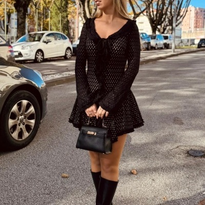 Mulher com vestido preto rendado, bolsa pequena preta e botas pretas em rua com árvores e carros.