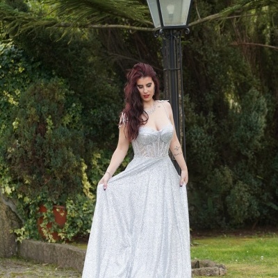 Mulher com vestido longo prateado brilhante em ambiente exterior com poste de luz e vegetação