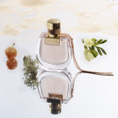 Frasco de perfume com líquido rosa claro e tampa dourada sobre superfície espelhada com ramos verdes, flor amarela e bagas vermelhas