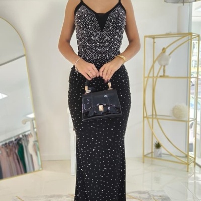 Mulher com vestido longo preto de bolinhas brancas e mala preta de mão num ambiente interior moderno.