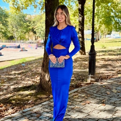 Mulher com vestido azul royal comprido com corte e nó, carteira azul brilhante e sapatos de salto bege numa calçada ao ar livre