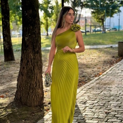 Mulher com vestido longo verde oliva pregueado e flor no ombro, segurando bolsa pequena, em passeio exterior com árvores.