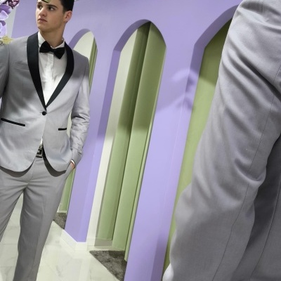 Homem com fato cinza claro olhando para o espelho numa sala lilás com flores artificiais