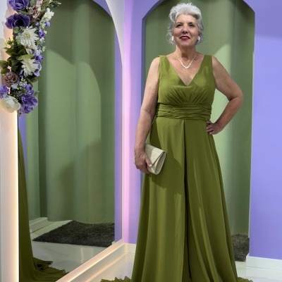 Mulher com vestido longo verde em ambiente com parede lilás, espelho e arco de flores artificiais.