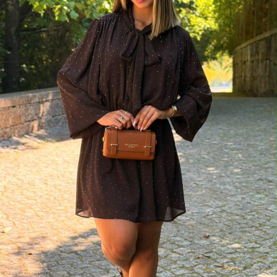 Mulher com vestido preto de bolinhas, sapatos pretos e carteira castanha, em passeio exterior com árvores.